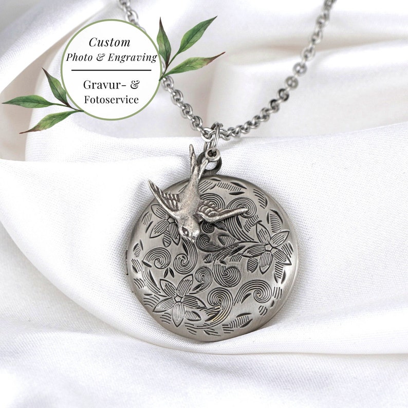 May include: A silver-toned locket necklace with a bird charm. The round locket features an engraved floral design and a small bird. The necklace includes the text "Custom Photo & Engraving" and "Gravur- & Fotoservice".