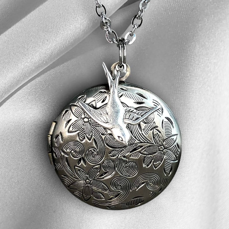 May include: Silver-toned locket necklace with a detailed floral pattern and a perched bird. The round pendant opens to reveal space for a photo or small keepsake. The necklace has a delicate chain.