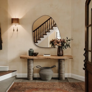 May include: A wooden console table with a live edge top and turned legs. The table is decorated with a vase of flowers, a ceramic vase, and a bowl. A large, organic-shaped mirror hangs on the wall behind the table.