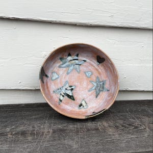 May include: A handmade ceramic bowl with a pink interior and a brown rim. The bowl is decorated with blue and black star and heart shapes. The bowl is sitting on a dark wooden surface.