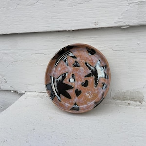 May include: A small, round ceramic dish with a speckled, light brown base. The dish is decorated with black and white abstract shapes, including stars and hearts. The rim is dark brown.