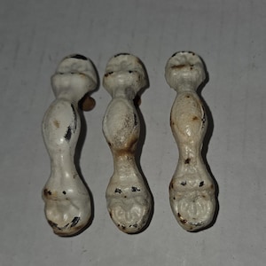 May include: Three antique white cast iron furniture feet with a distressed finish. Each foot has a rounded top, a bulbous midsection, and a hoof-like base. The feet show signs of age and wear, with visible rust and paint chipping.