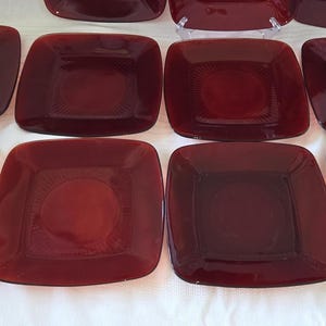 May include: A collection of square, deep red glass plates arranged in a grid. Each plate features a smooth, rounded square center surrounded by a textured border with radiating lines that extend toward the slightly raised, curved edges.
