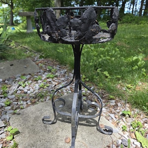 May include: A black metal plant stand featuring a circular top with a decorative leaf motif border. The stand is supported by three slender, curved legs that meet at a central point, providing a stable base for holding potted plants or decor.