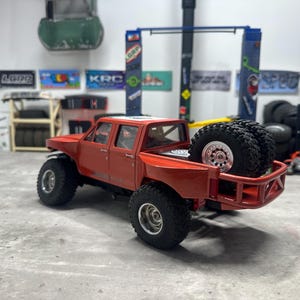 May include: A red toy pickup truck with large black tires and a roll cage. Two spare tires are mounted in the truck bed. The truck has a detailed interior and is set against a backdrop of workshop-themed decorations.