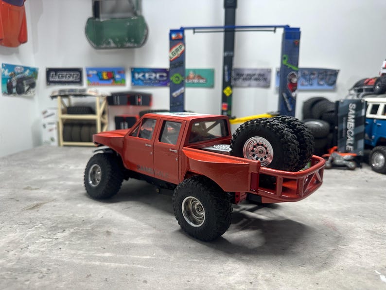 May include: A red toy pickup truck with large black tires and a roll cage. Two spare tires are mounted in the truck bed. The truck has a detailed interior and is set against a backdrop of workshop-themed decorations.