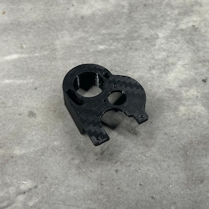 May include: A black, 3D-printed component with multiple circular openings and a textured surface. The object has a unique shape with a protruding section and several screw holes, suggesting a mechanical or technical application.