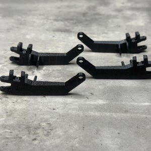 May include: A set of four black, 3D-printed mechanical components. Each piece features an elongated, angled arm with a circular mounting hole at one end and a complex, multi-pronged bracket structure at the opposite end for assembly.