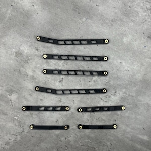 May include: A set of seven black, elongated, rigid components featuring a series of rectangular cutouts and small, circular brass-colored metal inserts at each end. These components vary in length and shape, with some featuring a slight angle.