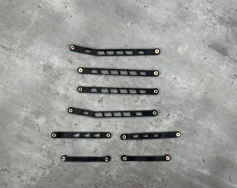 May include: A set of seven black, elongated, rigid components featuring a series of rectangular cutouts and small, circular brass-colored metal inserts at each end. These components vary in length and shape, with some featuring a slight angle.