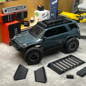 May include: A dark gray remote-controlled SUV model with black off-road tires, a roof rack, and a winch on the front bumper. The vehicle features the text "SAME SCALE" on the windshield and "LGRP" on the rear window. Additional black plastic roof rack components and side panels are displayed separately in the foreground.