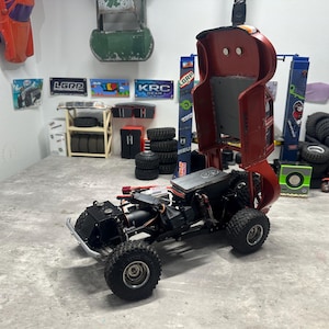May include: A partially assembled RC car chassis with large, knobby tires. The red body is raised, revealing the internal components. The scene includes various RC car parts and accessories, suggesting a hobbyist's workspace.