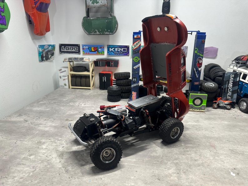 May include: A partially assembled RC car chassis with large, knobby tires. The red body is raised, revealing the internal components. The scene includes various RC car parts and accessories, suggesting a hobbyist's workspace.