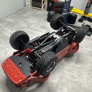 May include: A red and black remote control car chassis with large, knobby tires. The vehicle is upside down, showcasing its undercarriage and suspension system. The tires are black, and the frame is red.