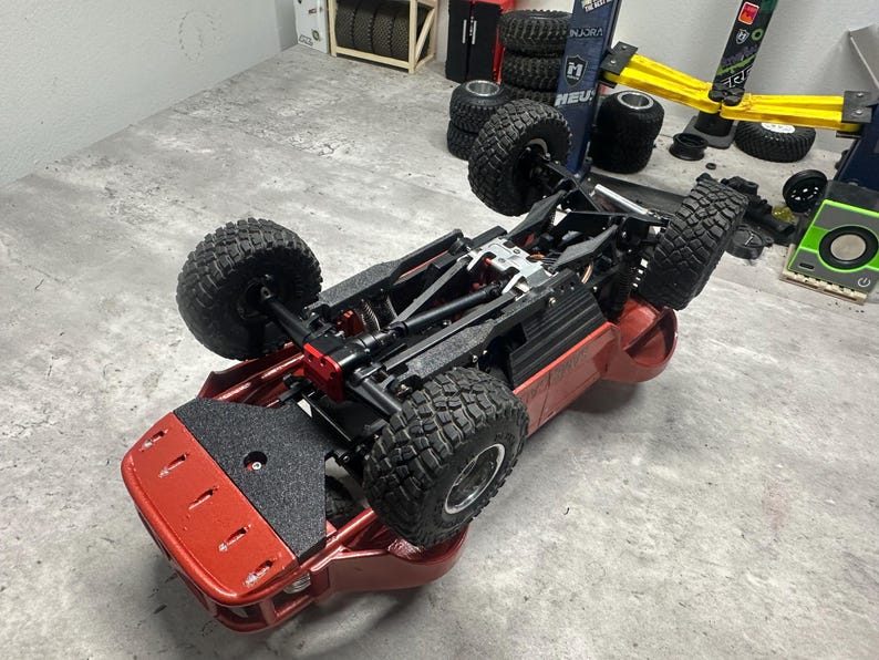 May include: A red and black remote control car chassis with large, knobby tires. The vehicle is upside down, showcasing its undercarriage and suspension system. The tires are black, and the frame is red.