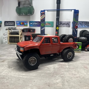 May include: A red toy pickup truck with large black tires. The truck has a detailed design, including a front bumper, side mirrors, and a bed. Additional tires are stacked in the truck bed. The background includes various signs and other toy vehicles.