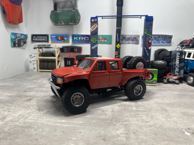 May include: A red toy pickup truck with large black tires. The truck has a detailed design, including a front bumper, side mirrors, and a bed. Additional tires are stacked in the truck bed. The background includes various signs and other toy vehicles.