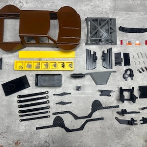 May include: A disassembled model car kit with various components. The brown body and roll cage are visible, along with gray and black parts, screws, and a yellow tray. The parts are arranged on a gray surface.