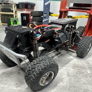 May include: A black and silver remote control truck with large, knobby tires. The truck has a silver front bumper and visible internal components, including wiring and suspension. The background includes tires, tools, and a red lift.