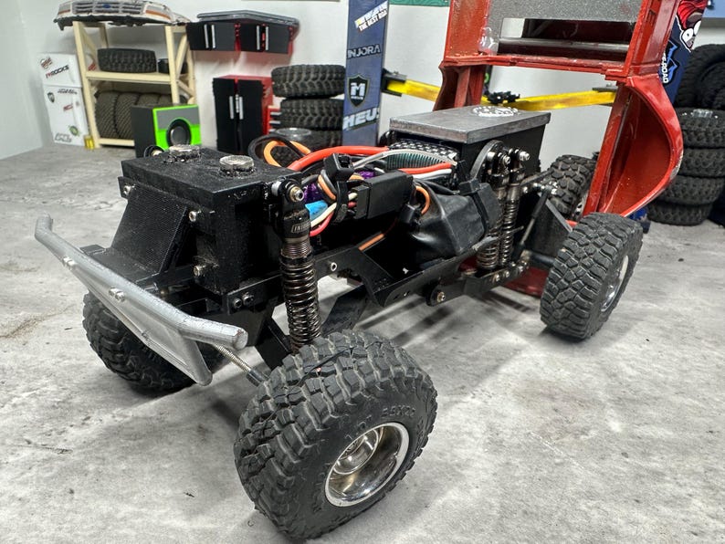 May include: A black and silver remote control truck with large, knobby tires. The truck has a silver front bumper and visible internal components, including wiring and suspension. The background includes tires, tools, and a red lift.