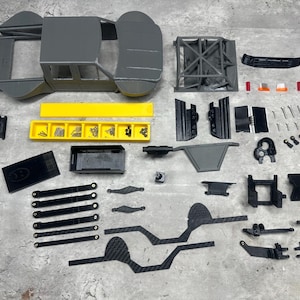 May include: An assortment of gray and black model car parts, including a chassis, roll cage, and various components. A yellow tray holds small hardware. The parts are arranged on a gray surface, suggesting a hobby or model-building project.
