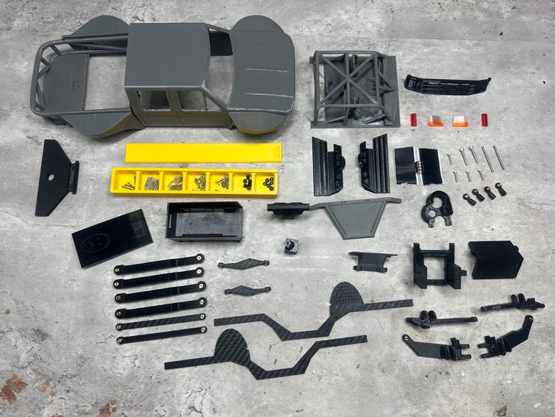 May include: An assortment of gray and black model car parts, including a chassis, roll cage, and various components. A yellow tray holds small hardware. The parts are arranged on a gray surface, suggesting a hobby or model-building project.