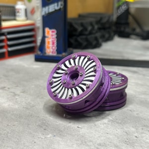 May include: Two purple and silver custom RC crawler wheels featuring a multi-spoke design with visible metal screws around the outer rim. The wheels have a layered construction with a deep purple outer ring and a silver inner turbine-style pattern.
