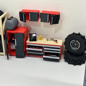 Puede incluir: Un pequeño cofre de herramientas rojo y negro con cajones, un armario rojo con puertas negras y dos armarios de pared rojos con puertas negras. El cofre de herramientas está sobre una superficie de madera con una pequeña caja, un martillo y una llave inglesa. También hay dos neumáticos negros con llantas plateadas.