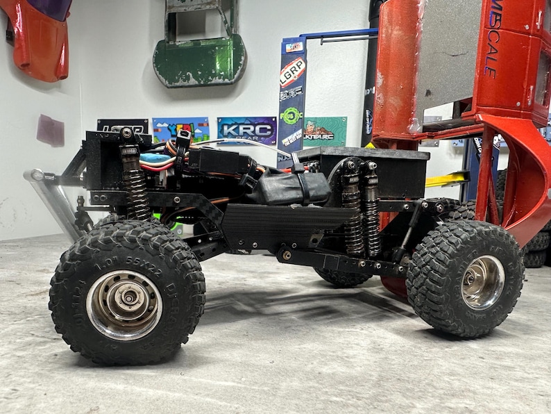 May include: Black and silver remote control truck with large, knobby tires. The truck has a black chassis, visible suspension, and a red body component. The tires are marked with "1.0" and "55x22".