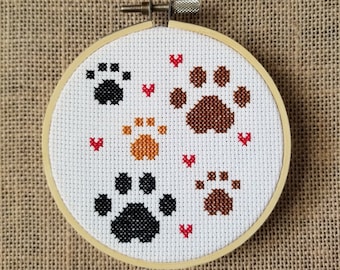 May include: A white embroidery hoop with a cross-stitch design of four paw prints in different colors: black, brown, orange, and red. The paw prints are surrounded by small red hearts.