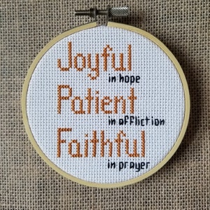 Könnte beinhalten: Ein weißer Stickrahmen mit den Worten "Joyful in hope, Patient in affliction, Faithful in prayer", die in orangem Garn gestickt sind.