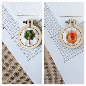 May include: Two small wooden embroidery hoops with cross-stitch designs. One features a green tree with red berries, the other a jar of jam with the word "JAM". A needle rests on the white fabric, with a grid pattern.