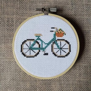 May include: A green bicycle with a basket of flowers on the handlebars. The bicycle is embroidered on white fabric and framed in a wooden hoop.