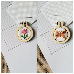 Puede incluir: Dos pequeños aros de bordado de madera con diseños de punto de cruz. El aro de la izquierda presenta un tulipán rosa con hojas verdes. El aro de la derecha presenta una mariposa marrón con alas blancas.