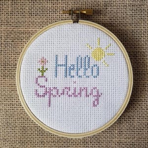 Puede incluir: Un aro de bordado blanco con las palabras "Hello Spring" bordadas en hilo rosa, azul y amarillo. Una pequeña flor está bordada a la izquierda del texto y un sol está bordado a la derecha.