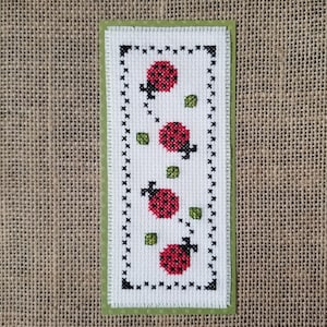 May include: A white cross-stitch bookmark with a green border and a black frame. The bookmark features four red ladybugs with black spots and green leaves.