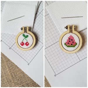 May include: Two small wooden embroidery hoops with white Aida cloth and cross-stitch designs. The hoop on the left has a red cherry design. The hoop on the right has a watermelon design.