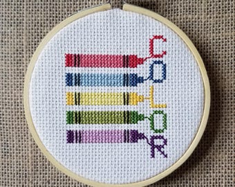 May include: A cross-stitch embroidery hoop featuring a design of colorful crayons and the word "COLOR" in a vertical orientation. The crayons are red, pink, blue, yellow, green, and purple. The embroidery is set against a white background.