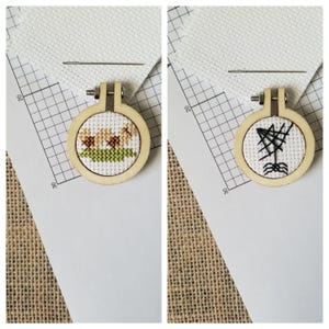 May include: Two small wooden embroidery hoops, each with a different cross-stitch design. One features brown and yellow flowers with green grass, the other a black spiderweb. A needle and white fabric are also visible.