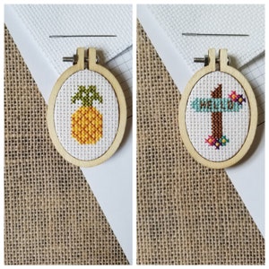 Puede incluir: Dos pequeños aros de madera para bordar con diseños de punto de cruz. El aro de la izquierda presenta una piña amarilla con hojas verdes. El aro de la derecha presenta un letrero marrón con la palabra "HELLO" en letras azules.