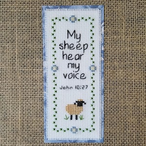 May include: White cross-stitch bookmark with a green border and blue flowers. The bookmark features a small brown sheep with black hooves and a black head. The text on the bookmark reads "My sheep hear my voice John 10:27".