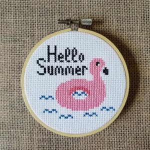 May include: A cross-stitch embroidery in a wooden hoop features a pink flamingo float with the words "Hello Summer" in black. Blue waves are stitched below the float. The background is a tan burlap.