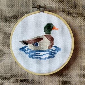 May include: A cross-stitch embroidery of a mallard duck swimming in a blue pond, framed in a natural wood hoop.