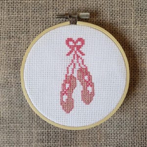 Puede incluir: Un bordado de punto de cruz rosa y blanco de un par de zapatillas de ballet atadas con un lazo, enmarcado en un aro de madera.