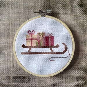 Puede incluir: Bordado de punto de cruz en un aro de madera que representa un trineo marrón que transporta tres regalos envueltos. Los regalos son en tonos beige, rosa y rojo, con lazos. El fondo es de tela de yute neutra.