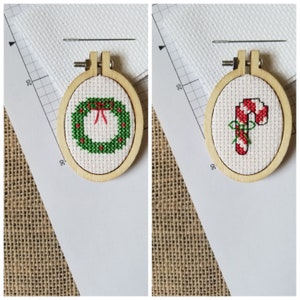 May include: Two small, wooden embroidery hoops each hold a cross-stitched Christmas design. One features a green wreath with a red bow, the other a red and white candy cane with a green bow. A needle rests above each hoop.