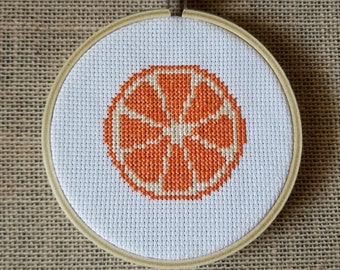Könnte beinhalten: Ein weißer Stickrahmen mit einem Kreuzstich-Design einer Orangenscheibe. Die Orangenscheibe ist leuchtend orange mit einem weißen Zentrum.