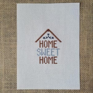 May include: A cross-stitch design on white fabric with the words "HOME SWEET HOME" in brown and blue thread. The design includes a brown house outline with decorative elements. The background is a burlap texture.