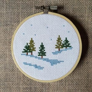 May include: A white cross-stitch embroidery hoop depicting a winter scene with three green trees and blue snow drifts. The background is a light blue sky with white stars.