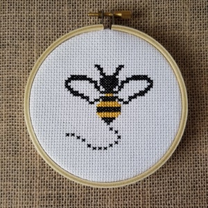 May include: A black and yellow bee cross-stitch embroidery in a round wooden hoop. The bee is flying with its wings spread and has a dotted line behind it.